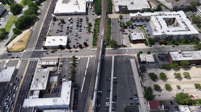 Flat commercial rooftops border a Rockville Centre rail corridor, Nassau County, Long Island, New York. The drone travels west, descending. Green foliage forms a textured pattern across the landscape.