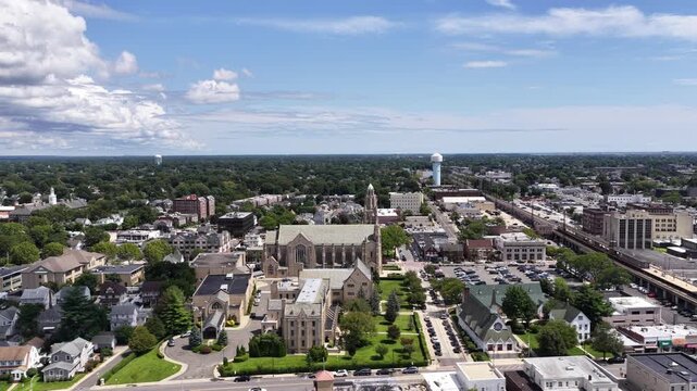 A church stands among streets and rooftops in Rockville Centre, Nassau County, Long Island, New York. The drone travels north while climbing. The landscape extends across the frame in summer daylight.