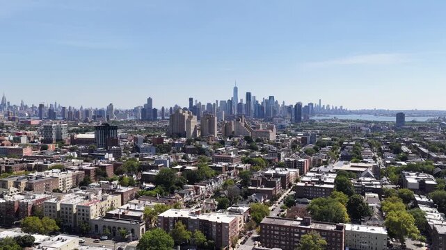 Jersey City rooftops lead toward the distant Manhattan skyline, Hudson County, New Jersey. The drone travels east at nearly level altitude. Green foliage forms a textured pattern across the landscape.