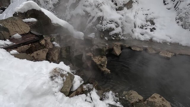 雪に覆われた地獄谷温泉の湯気が立ち上る岩風呂と源泉の湯口