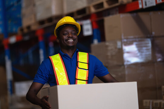 Smiling warehouse worker lifting a large cardboard box up from the floor in front of tall pallet racking, framed from a low angle in a logistics distribution centre