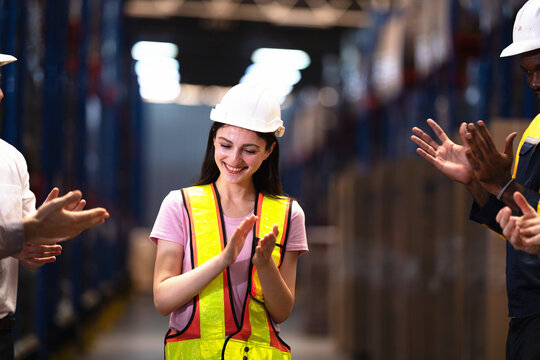 Smiling woman in a white hard hat and hi-vis vest claps her hands among applauding warehouse colleagues in a modern logistics distribution centre aisle in front of blue steel uprights