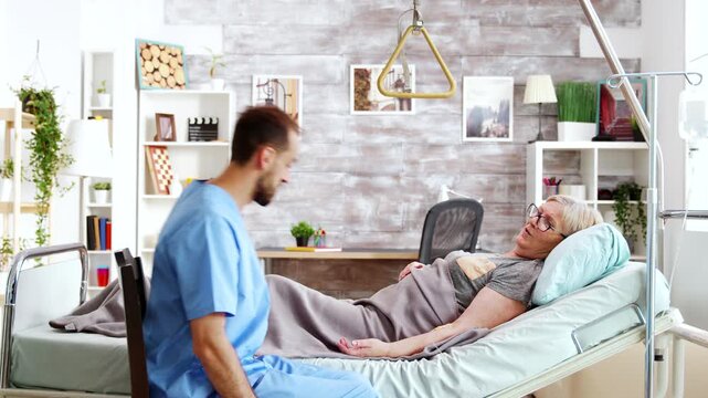 older woman smiles while talking with a male nurse beside her medical bed, conveying rapport, reassurance and supportive senior healthcare, further highlighting comfort and communication.