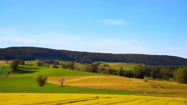 Rolling green and golden farmland drifts across frame during smooth lateral travel, revealing cultivated fields, scattered trees, wooded hills, and clear blue sky in bright daylight