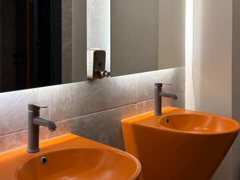 Two bright orange washbasins with modern faucets and illuminated mirror in a contemporary restroom interior with gray stone tiles and bold colorful design.