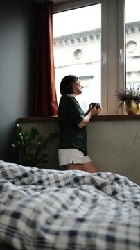 Woman stands by bedroom window with a mug, looking outside in a quiet morning moment, unmade bed nearby