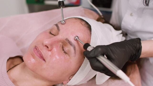 Female treatment. Skin therapy. Professional cosmetologist using modern aesthetic device for relaxing facial lifting procedure at workplace.