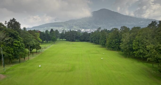 Aerial drone fly over golf course on Bali island. Golfers practicing their swing on lush green driving range, surrounded by tropical trees and distant mountains, enjoying outdoor leisure and sport