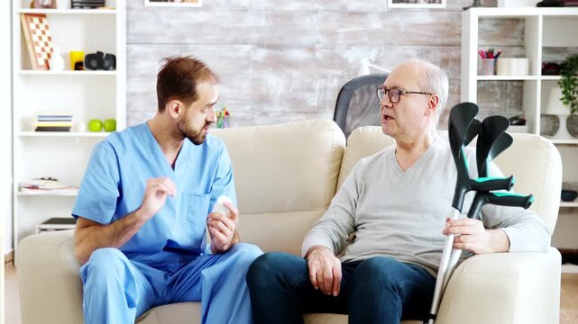 Male nurse discusses medication dosage with an older man beside his crutches, providing patient education, treatment guidance and attentive senior care, also emphasizing safety and mobility.