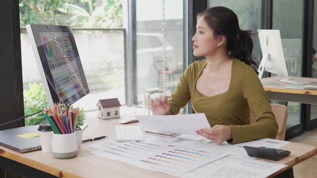 Young Asian female designer working on computer and taking notes on notebook in office