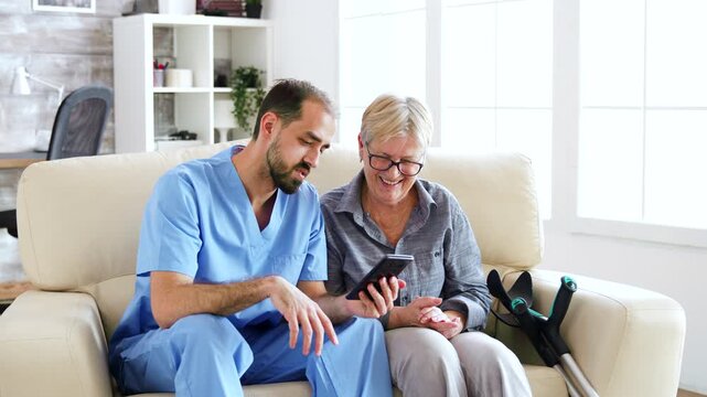 male nurse guides a senior woman using a smartphone while she sits beside crutches, highlighting digital inclusion, learning and supportive care.
