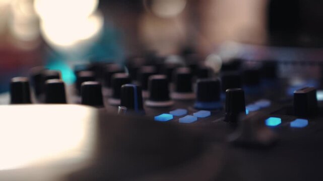 Close-up shot of a DJ hand adjusting knobs and mixing controls on an illuminated audio console during a live performance, vibrant LED lights glowing in a dark nightclub or festival environment