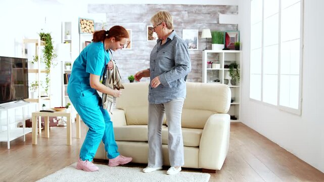 female nurse assists a senior woman with dressing beside the sofa, representing personal care, daily living support and dignified independence.