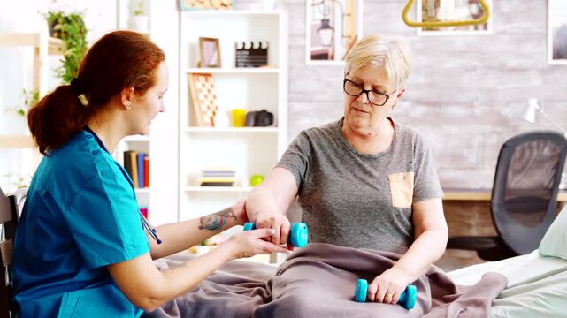 nurse supports an older woman performing seated dumbbell exercises on a care bed, illustrating strength training, rehabilitation and senior recovery support, further highlighting mobility.
