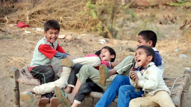 Small group of elementary age poor children sitting on charpai or cot outdoor in nature laughing video portrait together.