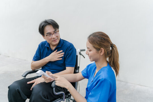 Nurse in blue scrubs checking temperature with infrared thermometer for senior male patient in wheelchair.