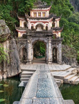 Ornate entrance gate of the Bich Dong Pagoda and Cave in Ninh Binh, Vietnam, featuring a stone path crossing a pond in front of traditional architecture and limestone mountain scenery
