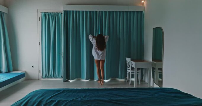 Woman opens blue curtains in a bright Bali hotel room balcony, arms outstretched toward mount Batur and a serene lake, enjoying tropical morning light, relaxation and travel freedom. Back view