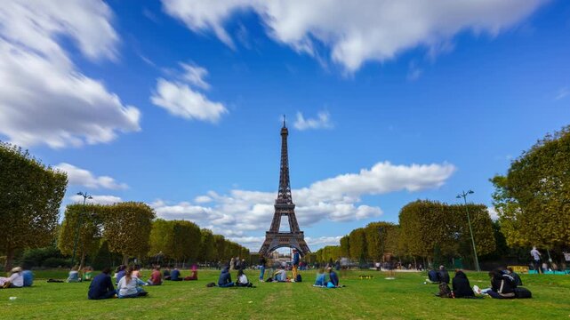 High-quality 4K cinematic timelapse footage of the iconic Eiffel Tower in Paris. Fluffy white clouds streak across a bright blue sky above the lush green lawns and autumn trees of the Champ de Mars.