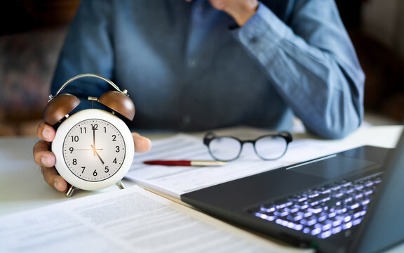 close up shot, asian businessman work in office with laptop and many documents he holding alarm clock at 5.00 PM end of work time in a day