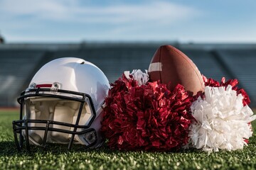 Obraz premium A white football helmet, brown football, and red and white pom-poms rest on green turf with stadium seats blurred in the background.