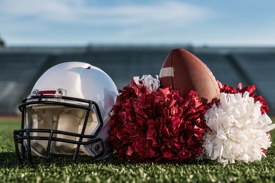 A white football helmet, brown football, and red and white pom-poms rest on green turf with stadium seats blurred in the background.