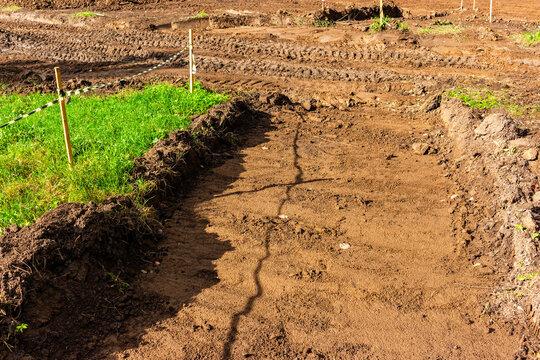 Heavy machinery tracks on disturbed brown soil during landscaping renovation in a city park, featuring temporary wooden stakes and barrier tape marking the dig site boundaries