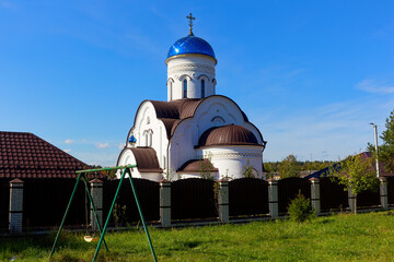 Stunning white orthodox church with blue domes stands proudly in russian village, rustic wooden swings sway gently on green lawn under bright blue sky © PhotoChur