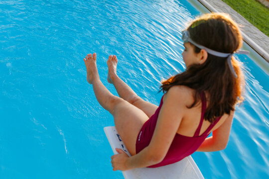 Young Girl Wearing Swimming Goggles Sitting on Diving Board Above Swimming Pool