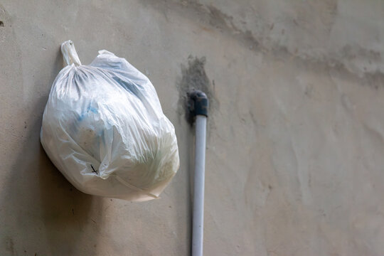White plastic trash bag hanging on concrete wall next to PVC pipe, waste disposal closeup