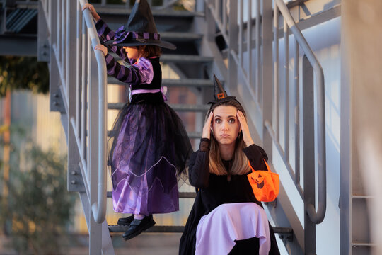 Mother Looking Tired Holding Head And Child In Witch Costume On Stairs For Halloween. Frustrated mother sitting on stairs holding pumpkin bag while her daughter climbs up in a witch costume