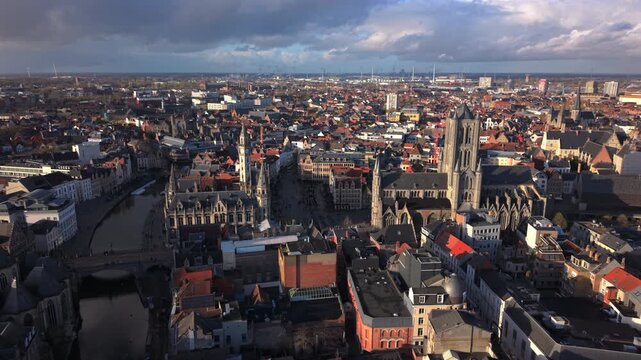 Cinematic aerial Ghent Belgium showcases stunning medieval cityscape with iconic Gothic towers rising above red rooftops. Historic canals wind through the old town center under dramatic cloudy skies.