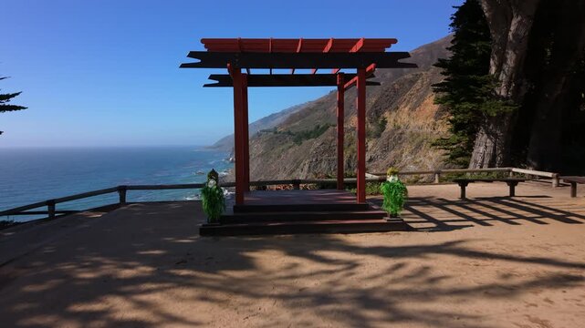 Empty wedding ceremony setup on coastal overlook featuring red wooden pergola altar centered at cliff edge and rows of white folding chairs aligned in foreground facing platform. Big Sur, California,