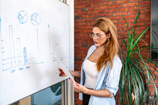 Business woman drawing charts on whiteboard. Female employee presenting financial data, corporate analysis, office presentation concept. Young manager looking at graphs and holding paper in hands.