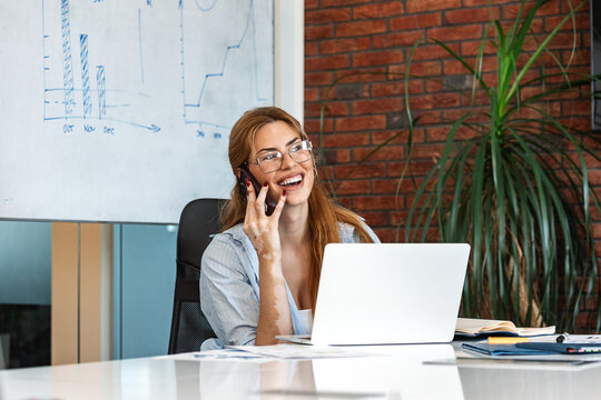 Business woman talking on smartphone in office. Female professional corporate phone call, startup communication concept. Smiling manager working on laptop computer at desk with whiteboards.