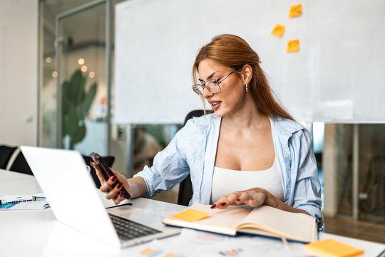 Business woman using smartphone and laptop in office. Female professional looking at mobile phone, corporate multitasking concept. Manager checking messages at modern workspace.
