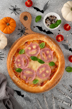Halloween pizza with smiling ham faces, basil leaves and spooky decorations on a gray background, creative seasonal food concept for parties, menus, and social media. Top view. Copy space.