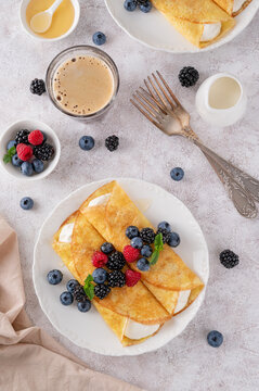 Crepes or thin pancakes with cream cheese, fresh berries and honey on a white plate on a concrete background. Copy space