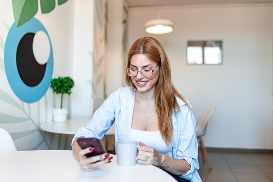 Business woman using smartphone and drinking coffee in modern office cafeteria. Smiling female professional reading mobile phone messages during break, relaxing in workplace lounge with eye wall art.