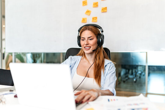 Business woman wearing headset with microphone working on laptop in office. Smiling female professional typing on computer, customer support, online call, remote work, customer service agent concept.