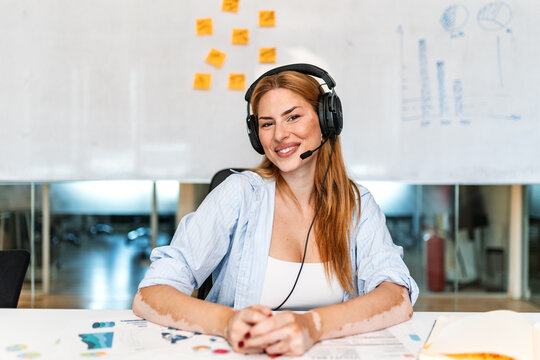 Business woman wearing headset with microphone sitting at desk in office. Smiling female professional looking at camera, customer support agent, call center operator, remote communication concept.
