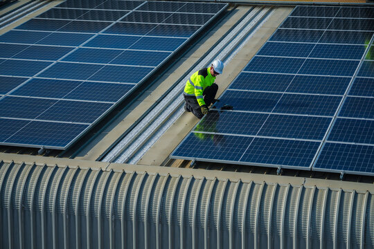 A photovoltaic technician wearing a safety vest and hard hat skilfully crouches over the panels for a final evening test on the sweeping metal roof of a manufacturing plant in Southeast.