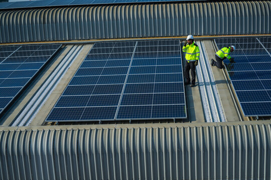 A photovoltaic technician wearing a safety vest and hard hat steadily crouches over the panels for a final evening test on the curved metal roof of an industrial factory in Bangkok Thailand.