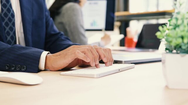 A close-up of a business professional typing on a white keyboard at a wooden desk in a modern office, with a laptop in the background and copy space.
