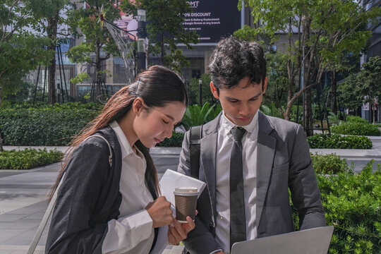 A young Asian businessman and his female colleague reviewing a file on a laptop held mid air at the foot of a glass skyscraper, illustrating leadership and partnership in modern business