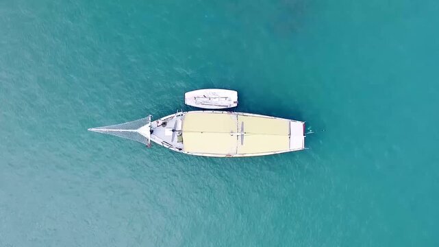 Aerial top-down view of a large sailboat with a small dinghy floating on calm turquoise ocean water