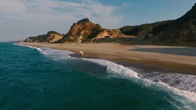 Aerial drone view of two people walking along a pebble beach beside turquoise ocean waves with coastal cliffs in the background
