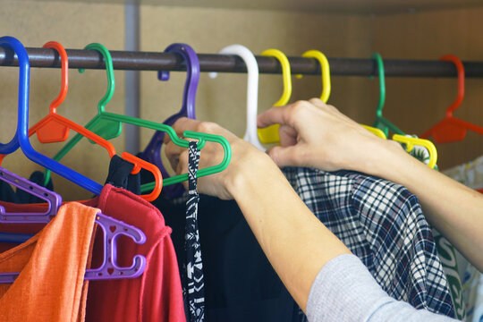 Woman's hands browsing plastic hangers with assorted clothing on rail inside wooden closet. Highlights everyday wardrobe choices, streamlined garment rotation, and organized apparel storage.