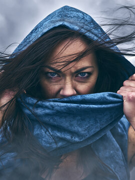 Photorealistic close-up of a woman pulling her blue hood and scarf over her face against strong wind
