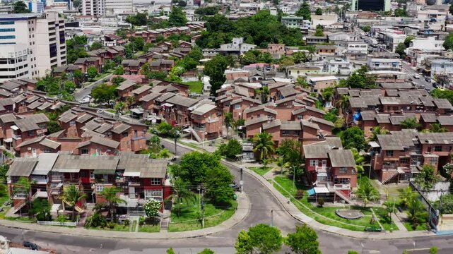 Aerial drone flies forward over rows of brick low rise social housing blocks of a conjunto habitacional by the igarape stream in Manaus, roundabout, parking and city towers behind, sunny day, Brazil.